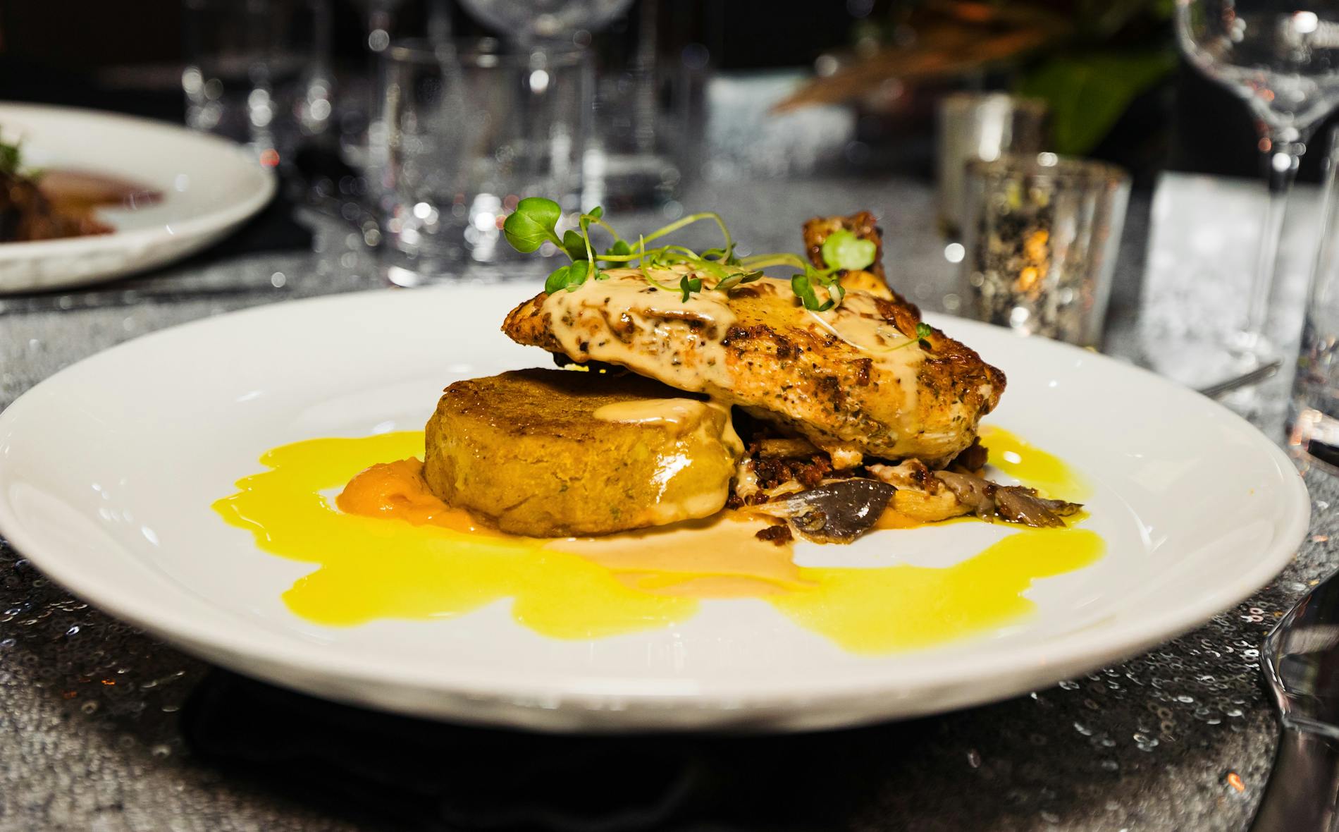 A beautifully plated Te Pae dish features a succulent piece of chicken atop a round, golden patty with a creamy sauce, all surrounded by vibrant sauce drizzles on a sparkling table setting.